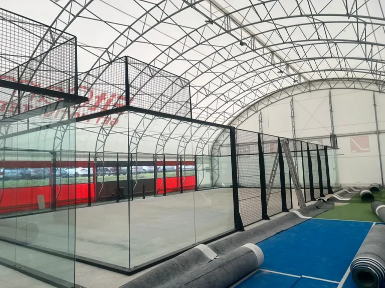 Sklenené steny a kovová mriežka dvoch padelových kurtov v hale FitCamp v Ružinove počas montáže, pri kurtoch rolky trávnika